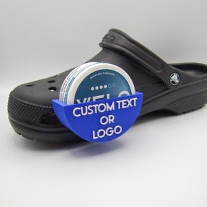 Puede incluir: Un zapato tipo zueco negro con un soporte azul que contiene un recipiente redondo. El soporte azul tiene el texto blanco "CUSTOM TEXT OR LOGO". El recipiente redondo tiene el texto "NICOTINE POUCHES".
