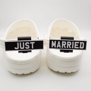 May include: Two white clogs with black and white text that reads "JUST" and "MARRIED". The text is on a black rectangular tag attached to the clog strap. The clogs are on a white surface.