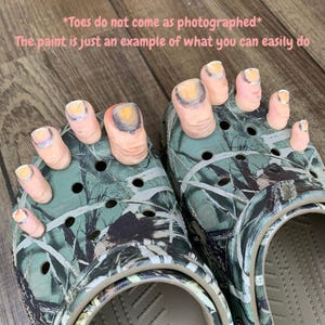 May include: A pair of camouflage Crocs with painted toe decorations. The toes are painted with a flesh-tone base and yellow and purple nail polish. Text on the image reads, "*Toes do not come as photographed* The paint is just an example of what you can easily do."