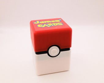 May include: A red and white card storage box with a black band and a white button. The top of the box is red with the words "James' CARDS" in yellow and blue.