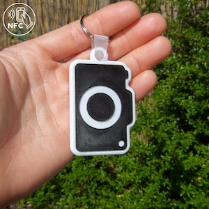 May include: A black and white keychain with a camera design. The keychain has a white border and a large central circle. The top has a clear plastic loop attached to a metal keyring. An NFC symbol is in the upper left corner.