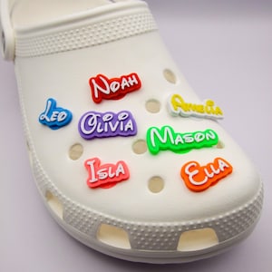 May include: White clog shoe adorned with colorful name charms. The charms feature names like "Noah," "Olivia," and "Ella" in various colors, including red, purple, and orange. The shoe has multiple ventilation holes.