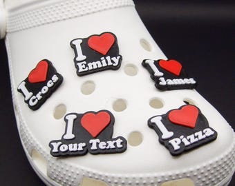 May include: A white foam clog shoe decorated with five black, white, and red plastic charms. Each charm features a red heart and the text I Crocs, I Emily, I James, I Your Text, and I Pizza, showcasing personalized designs for footwear accessories.