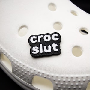 Könnte beinhalten: Schwarz-weißer "CROC SLUT"-Anhänger für ein Paar Crocs-Schuhe.