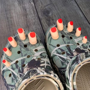 May include: A pair of camouflage Crocs adorned with prosthetic toes. Each toe is painted with a bright red nail polish. The shoes are designed to resemble feet.