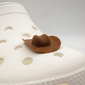 May include: A brown cowboy hat shoe charm rests on a white shoe. The hat features a textured brim and crown, offering a Western aesthetic. The shoe has several holes.