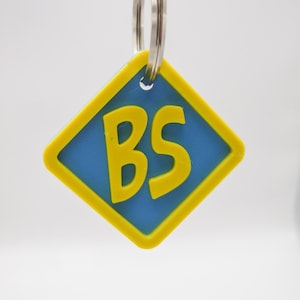 May include: A diamond-shaped keyring in yellow and blue, featuring the yellow letters "BS". The keyring is attached to a silver metal ring. The background is plain white.