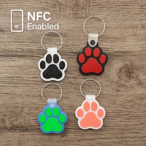 Puede incluir: Cuatro llaveros con forma de huella de pata con diferentes colores de huellas. Las huellas son negras, rojas, verdes y naranjas. Los llaveros están hechos de plástico blanco y tienen un anillo de metal plateado. El texto "NFC Enabled" es visible en la esquina superior izquierda de la imagen.