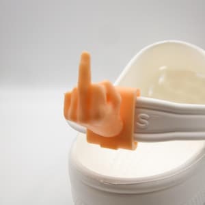 May include: A light orange hand gesture charm, showing the middle finger, attached to a white shoe. The charm is a detailed miniature of a hand. The shoe has a white strap and a white base.