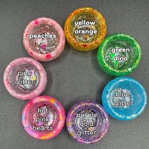 May include: Six circular resin bowls arranged in a circle. The bowls are labeled: peaches, yellow orange, green dino, blue glitter, purple gold glitter, and hot pink hearts. Each bowl features distinct colors, glitter, and embedded decorative elements.