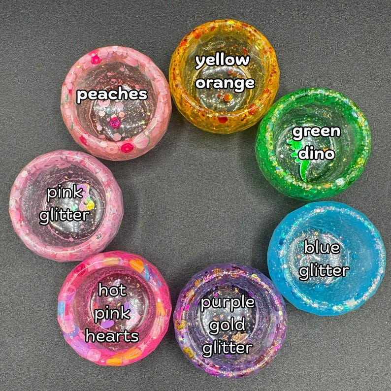 May include: Six circular resin bowls arranged in a circle. The bowls are labeled: peaches, yellow orange, green dino, blue glitter, purple gold glitter, and hot pink hearts. Each bowl features distinct colors, glitter, and embedded decorative elements.