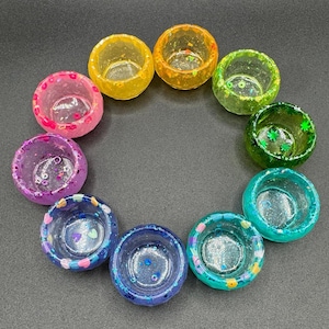 May include: A collection of ten small, circular resin bowls arranged in a circle. Each bowl features a unique color, including yellow, orange, light green, dark green, teal, blue, purple, and pink, with embedded glitter and small decorative shapes.