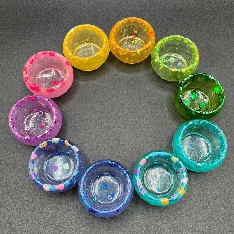 May include: A collection of ten small, circular resin bowls arranged in a circle. Each bowl features a unique color, including yellow, orange, light green, dark green, teal, blue, purple, and pink, with embedded glitter and small decorative shapes.