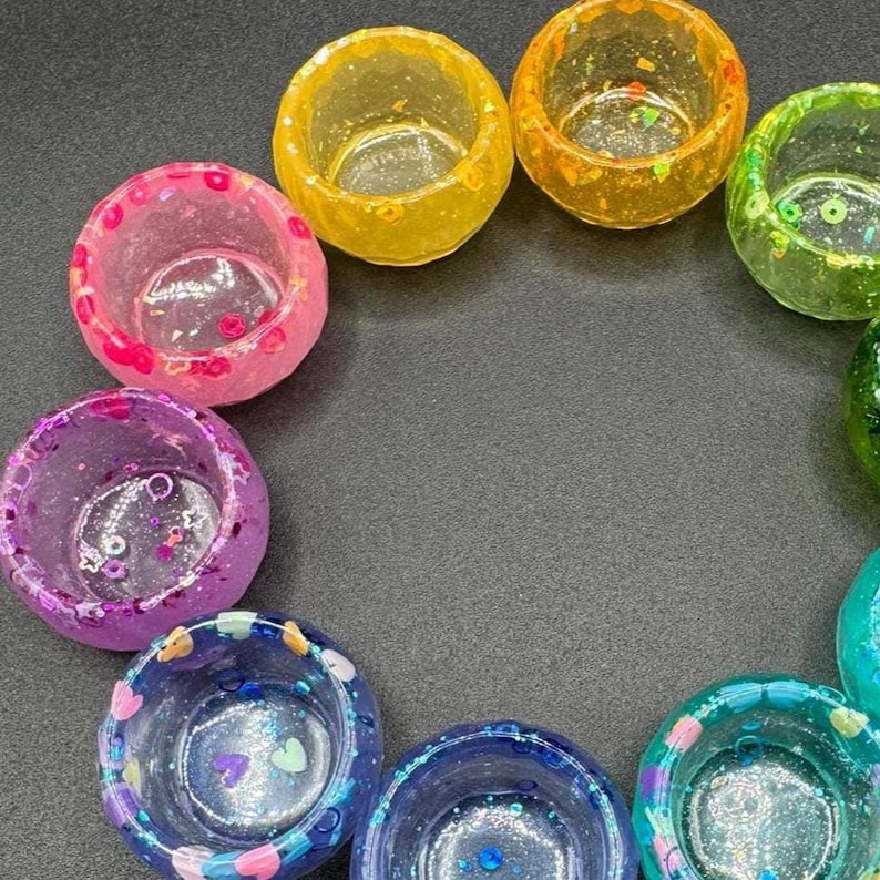 May include: A collection of small, circular resin bowls arranged in a circular pattern. Each bowl features a translucent finish with embedded colorful glitter and small decorative shapes in shades of yellow, orange, green, pink, purple, and blue.