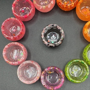 May include: A collection of small, circular resin bowls in various colors including pink, orange, green, and black. The bowls feature embedded glitter, sequins, and small decorative shapes. One black bowl in the center displays a white ghost graphic.