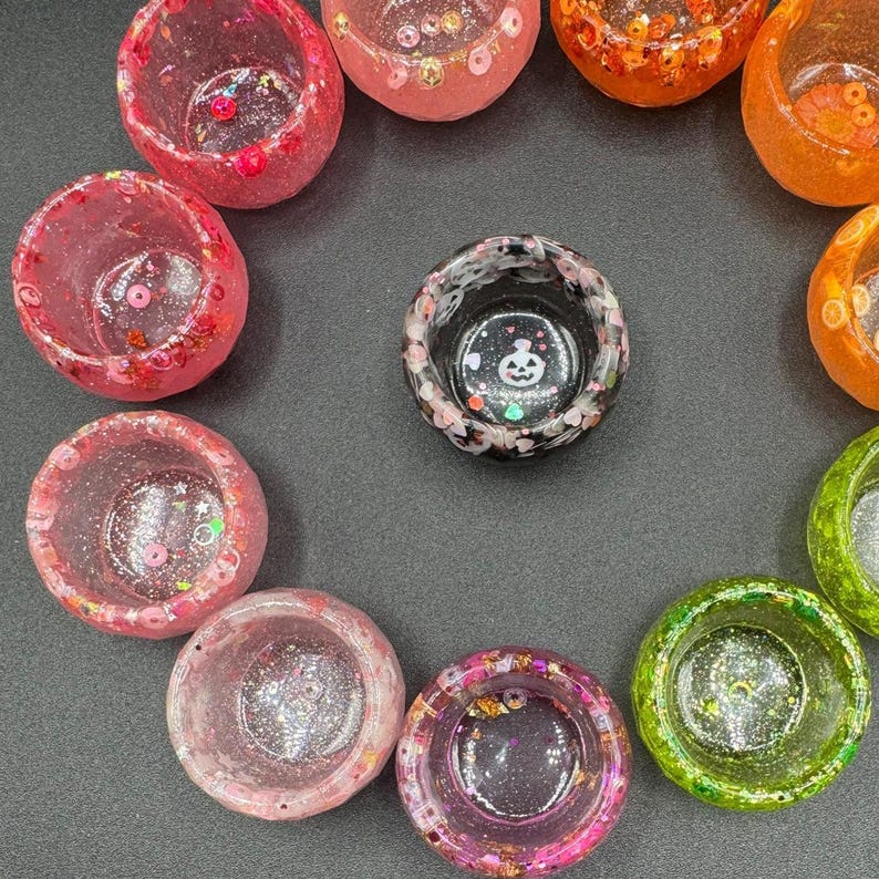 May include: A collection of small, circular resin bowls in various colors including pink, orange, green, and black. The bowls feature embedded glitter, sequins, and small decorative shapes. One black bowl in the center displays a white ghost graphic.