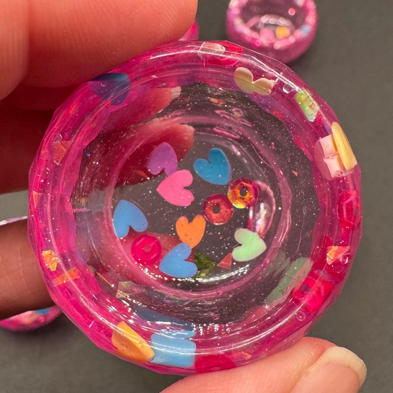 May include: A small, circular, translucent pink resin bowl featuring embedded colorful heart-shaped charms, small round beads, and iridescent glitter flakes. The bowl has a thick, textured rim and a clear, glossy finish, showcasing a variety of multi-colored decorative elements suspended within the clear resin material.