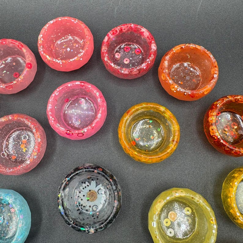 May include: A collection of ten small, circular resin bowls in various colors including pink, orange, yellow, and black. Each bowl features a translucent finish embedded with decorative elements like glitter, sequins, and small floral shapes.