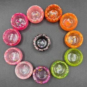 May include: A collection of ten small, circular resin bowls arranged in a circle, featuring various colors including pink, orange, green, and clear, with embedded glitter and confetti, plus one central black bowl with a white ghost graphic.