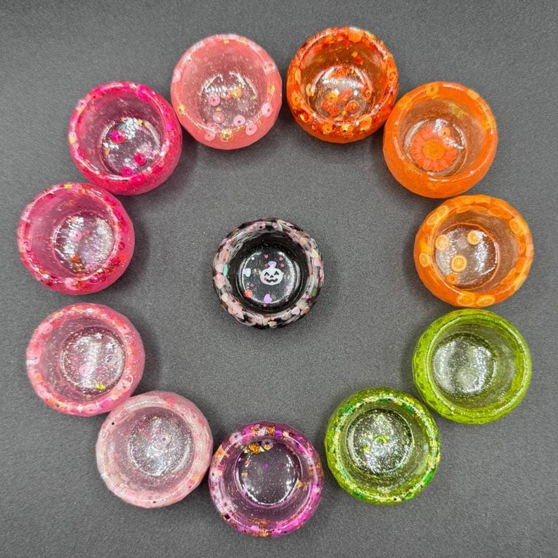 May include: A collection of ten small, circular resin bowls arranged in a circle, featuring various colors including pink, orange, green, and clear, with embedded glitter and confetti, plus one central black bowl with a white ghost graphic.