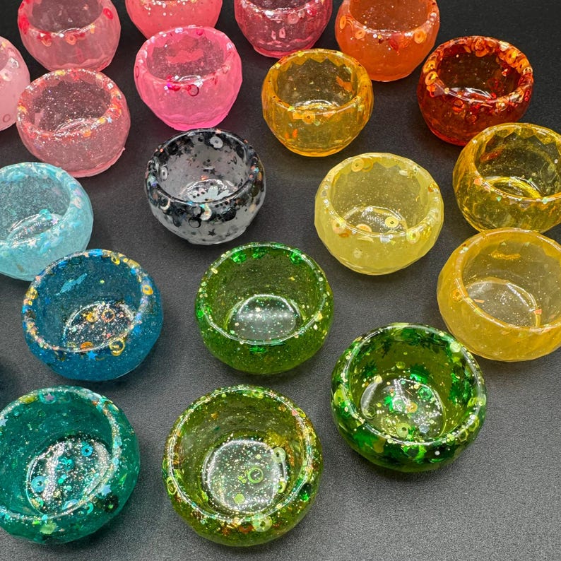 May include: A collection of small, round, translucent resin bowls in various colors including pink, orange, yellow, green, blue, and black. Each bowl features embedded glitter, sequins, and small decorative shapes throughout the material.