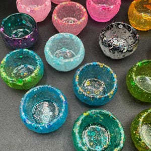 May include: A collection of small, round, bowl-shaped resin containers in various translucent colors including pink, blue, green, yellow, and black. Each container features embedded glitter, sequins, and star-shaped accents throughout the material.