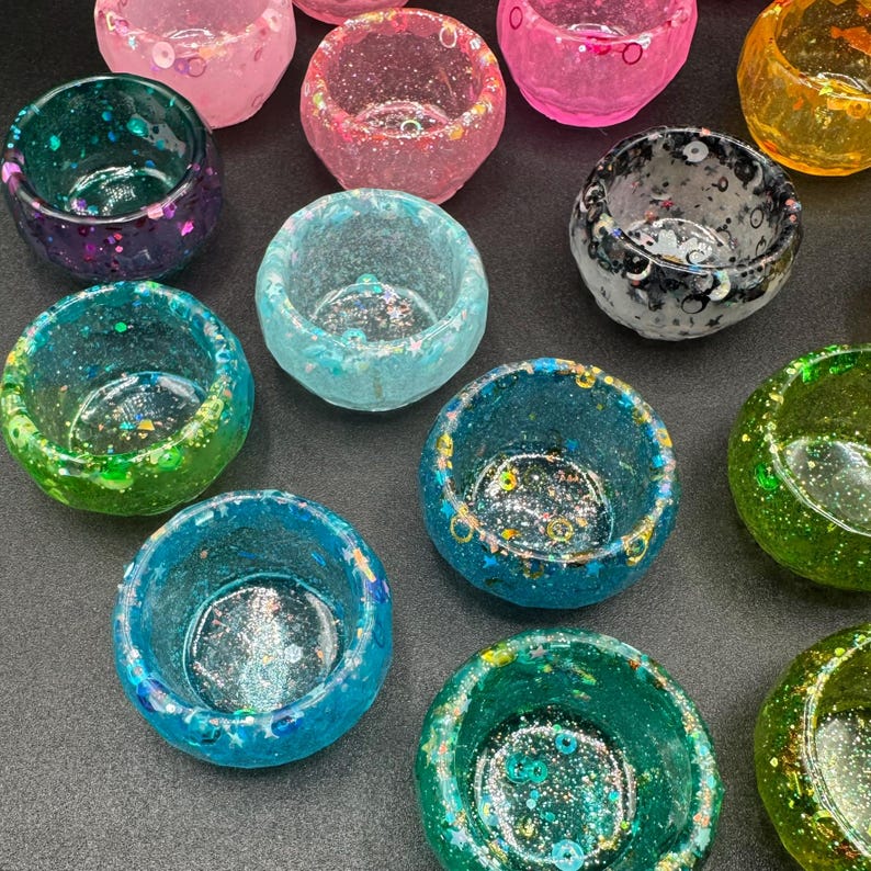 May include: A collection of small, round, bowl-shaped resin containers in various translucent colors including pink, blue, green, yellow, and black. Each container features embedded glitter, sequins, and star-shaped accents throughout the material.