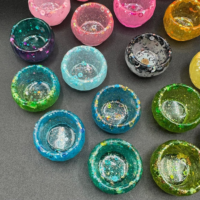 May include: A collection of small, round, bowl-shaped containers made of translucent resin. Each bowl features a unique color, including shades of pink, green, blue, yellow, and black, with embedded glitter, sequins, and decorative flecks throughout.