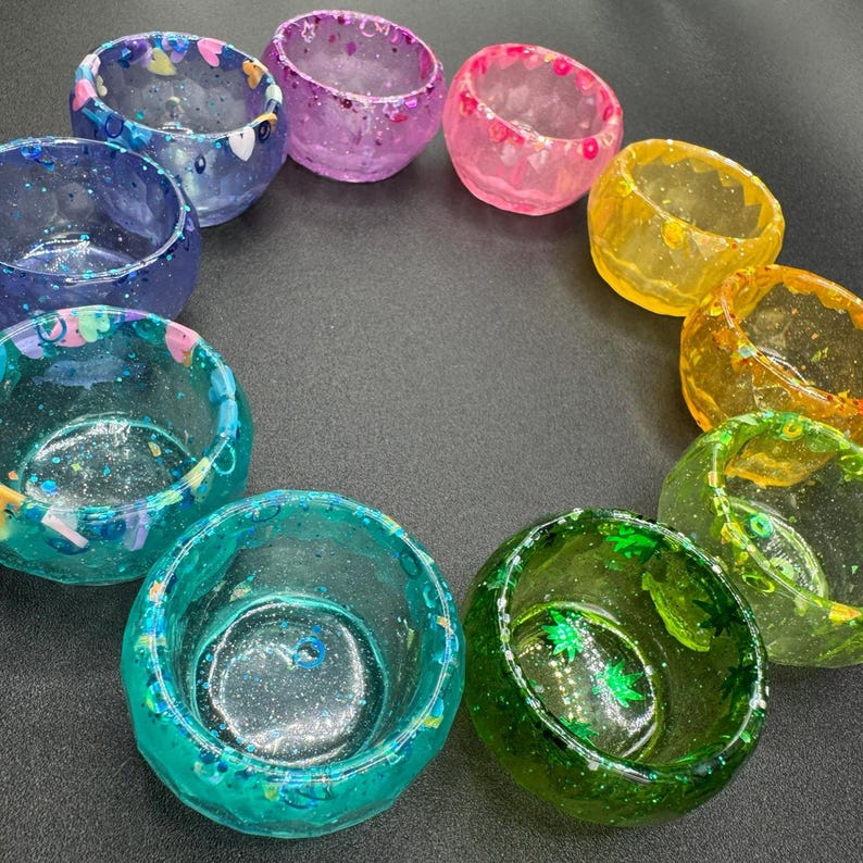 May include: A collection of ten small, circular resin bowls arranged in a circle. Each bowl features a unique color, including shades of blue, purple, pink, yellow, orange, and green, with embedded glitter and various small decorative shapes.