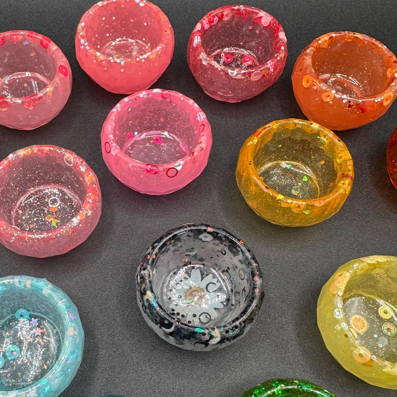 May include: A collection of small, round, translucent resin bowls in various colors including pink, orange, yellow, blue, and black. Each bowl features embedded decorative elements like glitter, small sequins, and tiny fruit-shaped slices.