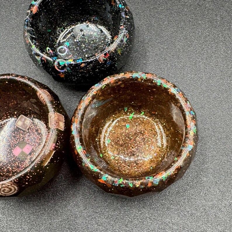 May include: Three small, round, bowl-shaped containers with a smooth, glossy finish. One container is black with iridescent glitter, while the other two feature dark brown tones with embedded metallic sparkles and small, colorful square accents.