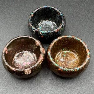 May include: Three small, round, bowl-shaped containers with faceted outer surfaces. One is black with multicolored glitter, one is dark brown with pink square accents and swirls, and one is a warm brown with green and orange flecked inclusions.