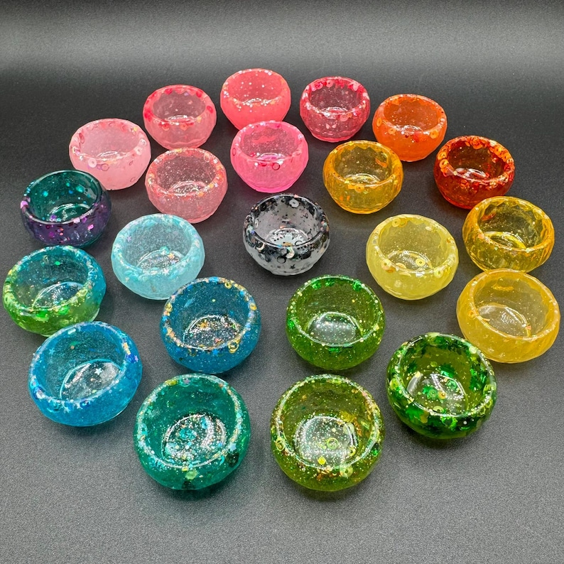 May include: A collection of twenty-one small, round, bowl-shaped resin containers arranged in a cluster. Each container features a unique translucent color, including shades of pink, orange, yellow, green, blue, and purple, all embedded with glitter.