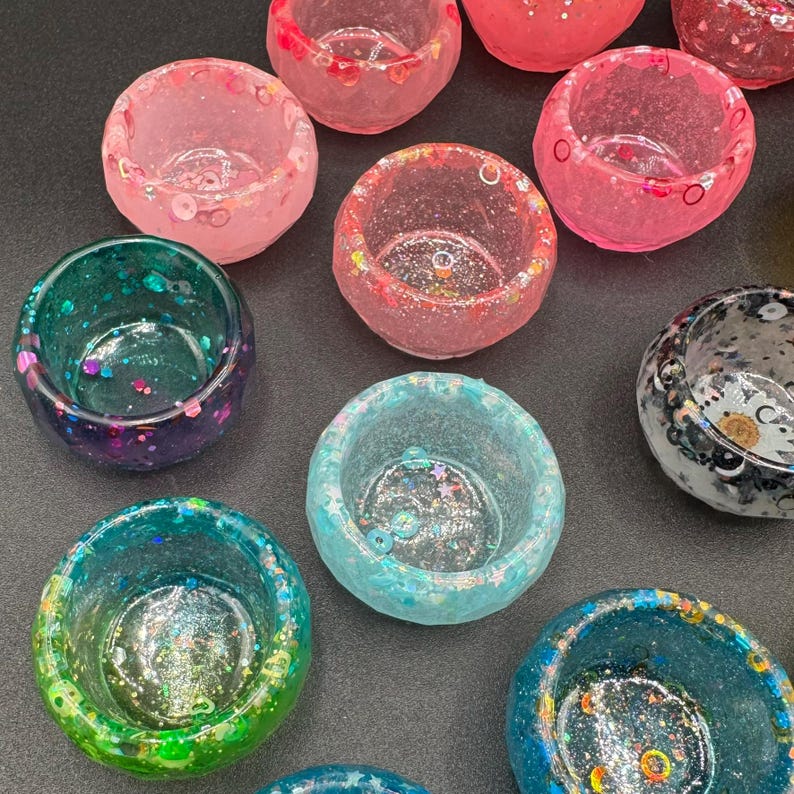 May include: A collection of small, round, bowl-shaped containers made of translucent resin. Each bowl features embedded glitter, sequins, and small decorative shapes in various colors including pink, teal, green, blue, and black with white speckles.