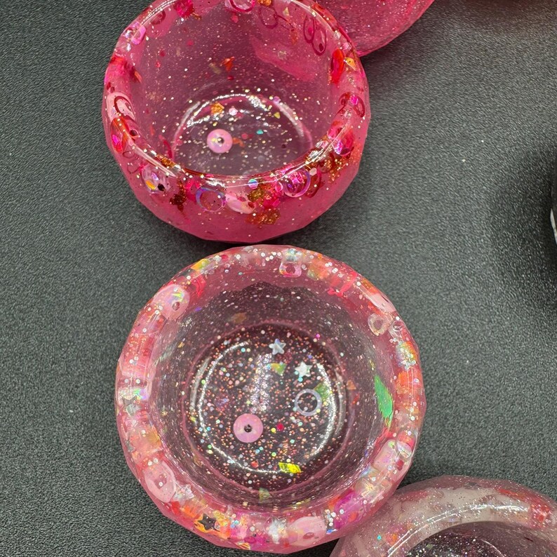 May include: Two circular resin bowls in a vibrant pink hue, featuring a translucent finish embedded with various pink-toned glitter, small star-shaped sequins, and circular confetti pieces throughout the thick, rounded walls and base.