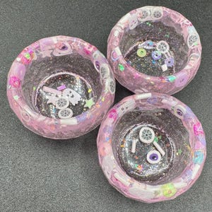 May include: Three round, translucent pink resin bowls with a glittery finish. The bowls are embedded with small, colorful decorative charms including white ghosts, purple and white eyeballs, stars, and circular beads throughout the material.