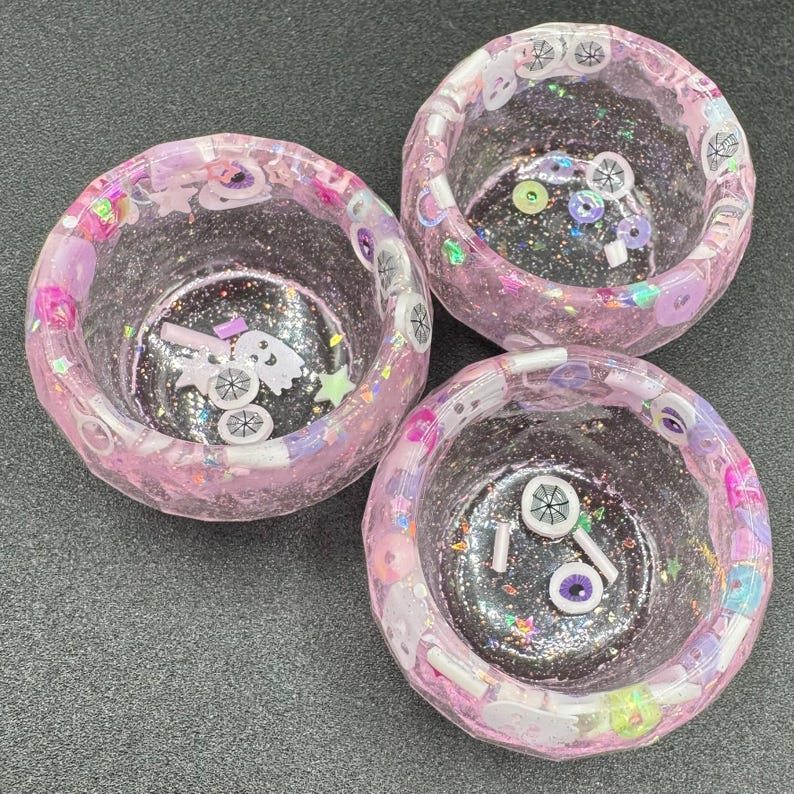 May include: Three round, translucent pink resin bowls with a glittery finish. The bowls are embedded with small, colorful decorative charms including white ghosts, purple and white eyeballs, stars, and circular beads throughout the material.