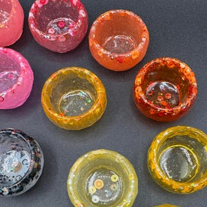 May include: A collection of small, round, translucent resin bowls in various colors including pink, orange, red, yellow, and black. Each bowl features embedded decorative elements like glitter, sequins, and small fruit-shaped slices.