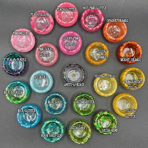 May include: A collection of twenty-one small, circular resin bowls arranged in a cluster. Each bowl features distinct colors and textures, including glitter, sequins, and translucent finishes, with text labels identifying each specific style.