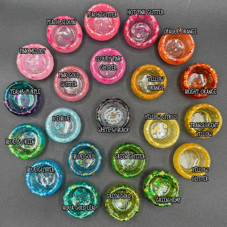 May include: A collection of twenty-one small, circular resin bowls arranged in a cluster. Each bowl features distinct colors and textures, including glitter, sequins, and translucent finishes, with text labels identifying each specific style.