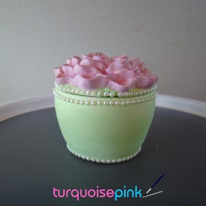 Puede incluir: Una caja de baratijas redonda de color verde claro con una tapa floral rosa. La caja está decorada con una hilera de perlas falsas en la parte superior e inferior. El texto "turquoise pink" está en la parte inferior.