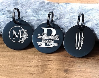 Könnte beinhalten: Drei schwarze Schlüsselanhänger aus Metall mit gravierten Initialen und Blumendesigns. Die Schlüsselanhänger zeigen die Buchstaben M, B und W, wobei der mittlere Schlüsselanhänger auch den Namen "Bonnie" enthält.