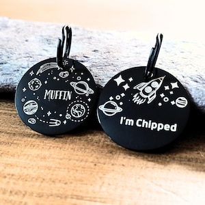 May include: Two black round pet ID tags with silver rings. The first tag has a space theme with planets, stars, and a rocket ship. The text "MUFFIN" is engraved on the tag. The second tag has the same space theme and the text "I'm Chipped" is engraved on the tag.
