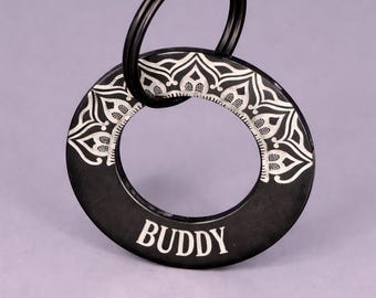 Puede incluir: Etiqueta negra para mascotas con el nombre "BUDDY" en letras blancas. La etiqueta tiene un diseño circular con un patrón floral blanco decorativo en el borde. Un anillo negro está adjunto.