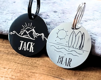 May include: Two round black and silver dog tags with key rings. The black tag has a mountain scene with a sun and the word "JACK" engraved on it. The silver tag has a beach scene with a sun, waves, and surfboards and the word "BEAR" engraved on it.