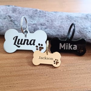 May include: Three bone-shaped dog tags with engraved names. The tags are silver, gold, and black. The names are "Luna", "Mika", and "Jackson".
