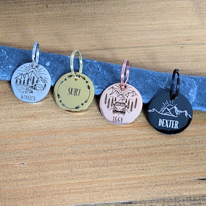 May include: Four round pet tags with silver, gold, and rose gold finishes. Each tag features a unique design: a mountain scene with a tent, a wreath with text, a camper van in a forest, and a mountain scene. The tags are engraved with the names Winston, Suri, Iggy, and Dexter.