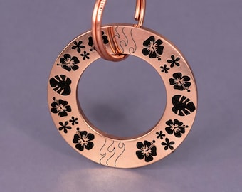 Puede incluir: Un llavero de color cobre con un colgante circular. El colgante está decorado con diseños florales y de hojas negras. Un anillo de color cobre está unido a la parte superior del colgante.