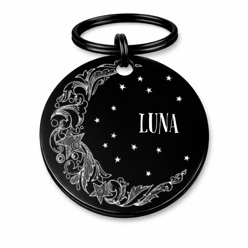 May include: Black circular pet tag with a decorative crescent moon and star design. The word "LUNA" is printed in white. A black metal ring is attached at the top for easy attachment to a collar.