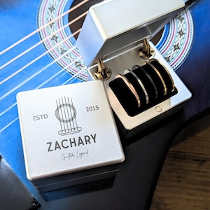 May include: A silver box with a hinged lid, open to reveal four bracelets in gold, silver, and black. A smaller square box with the text "ZACHARY Guitar Legend" and a guitar graphic sits in front. The boxes rest on a blue acoustic guitar.